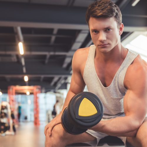 Attractive young muscular man is working out with dumbbells in gym