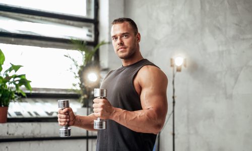 Powerful guy with muscular hands and huge biceps posing with dumbells in vintage studio room with special illumination.