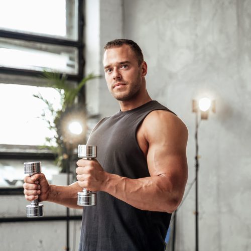 Powerful guy with muscular hands and huge biceps posing with dumbells in vintage studio room with special illumination.