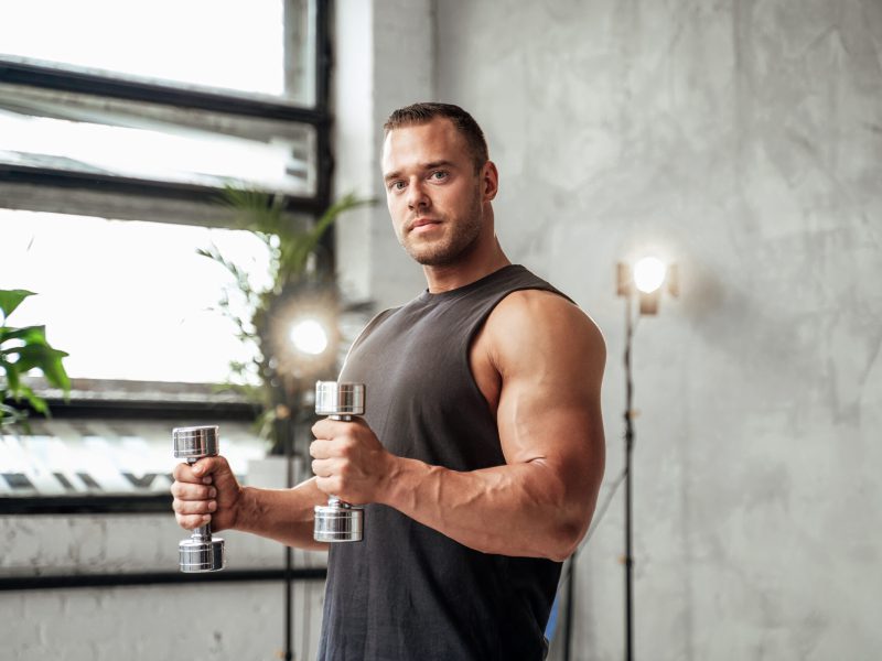 Powerful guy with muscular hands and huge biceps posing with dumbells in vintage studio room with special illumination.